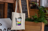 A cream cotton tote bag with the Crush ICE design -- a raised fist in progress pride colours crushing a chunk of ice -- hanging from a wooden furniture stand, with a fern planter and café chalkboard in the background.