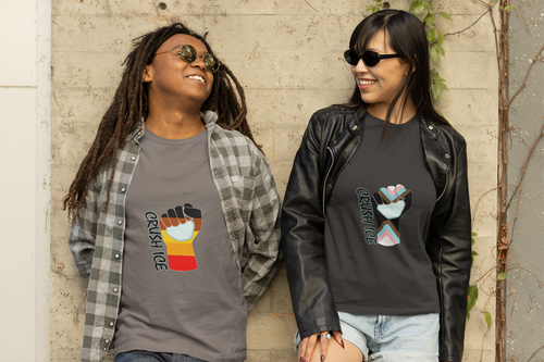 Two people of color stand in solidarity against an urban wall. On the left, a Black individual wears a charcoal grey unisex tee with the Crush ICE BIPOC Solidarity design—a brown and black protest fist shattering ice. On the right, a person with dark hair wears a black tee with the Transgender Legacy design, featuring a blue, pink, and white chevron fist. Both wear streetwear and sunglasses, representing intersectional resistance and community power.