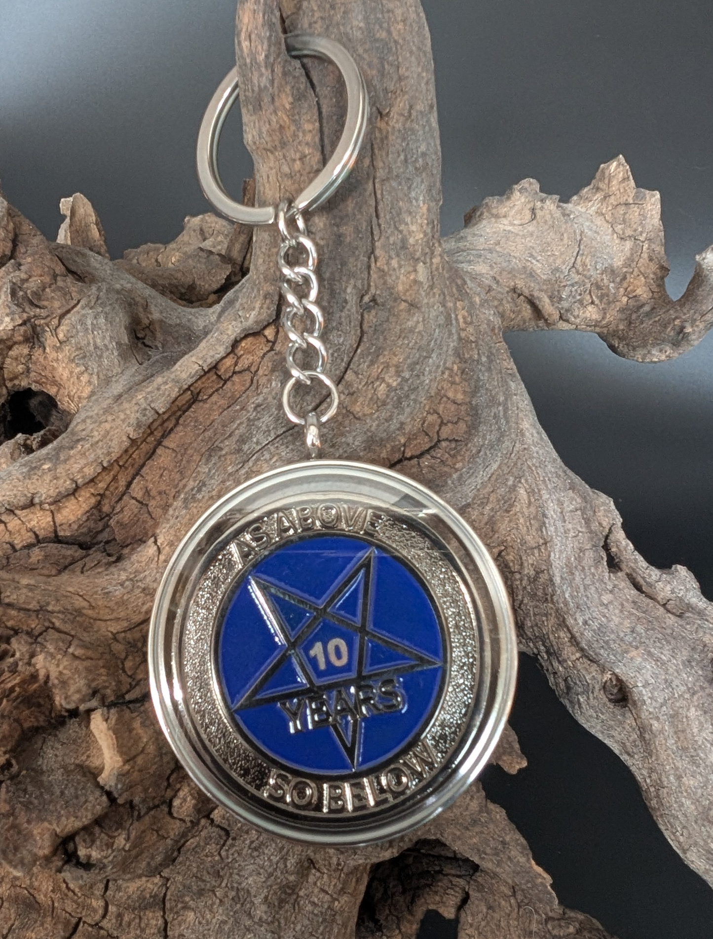 A keychain hangs from a piece of driftwood. The split ring is of the flattened style, with a short chain connected to a coin case. The case has steel sides that are screwed together and the faces are clear acrylic. Inside the case is a sobriety coin showing 10 years.