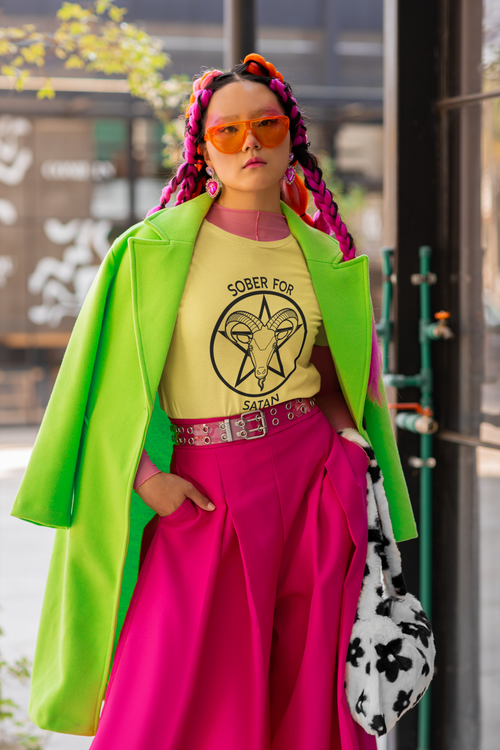 Photo of a fem-presenting person wearing a Bella Canvas round-neck t-shirt and a bold neon outfit as she poses in the street. The Tshirt is yellow, with a black Sober for Satan graphic on the front.