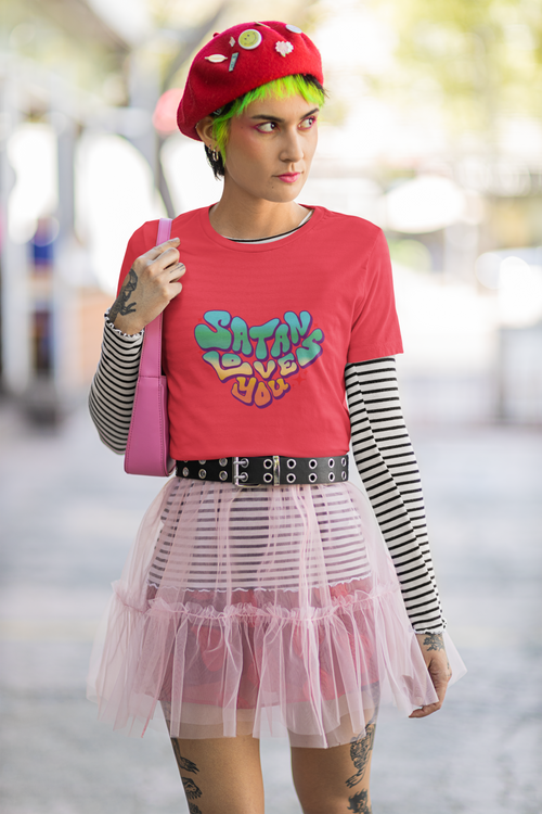 A fem-presenting person is wearing colourful, quirky outfit and a Bella Canvas t-shirt while posing in the street with a serious look. The tshirt has a groovy Satan Loves You graphic on the front.