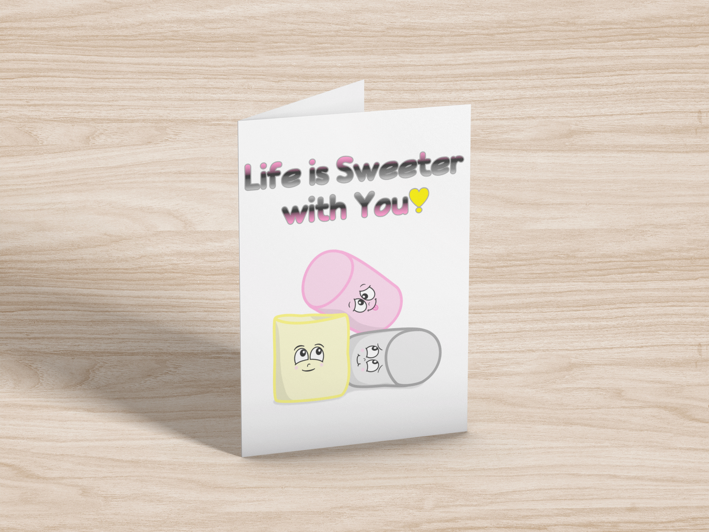 A greeting card on light wood background. The card has a white background. At the top is the text, "Life is Sweeter with You!" in the queerplatonic pride flag colours. Below are matching pink, yellow, and grey cartoon marshmallows with cute expressions.