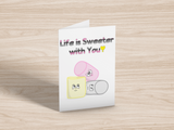 A greeting card on light wood background. The card has a white background. At the top is the text, "Life is Sweeter with You!" in the queerplatonic pride flag colours. Below are matching pink, yellow, and grey cartoon marshmallows with cute expressions.