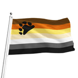 A waving bear pride flag on a silver pole, featuring horizontal stripes in brown, orange, pale yellow, white, gray, and black, with a large black paw print on the upper left side.