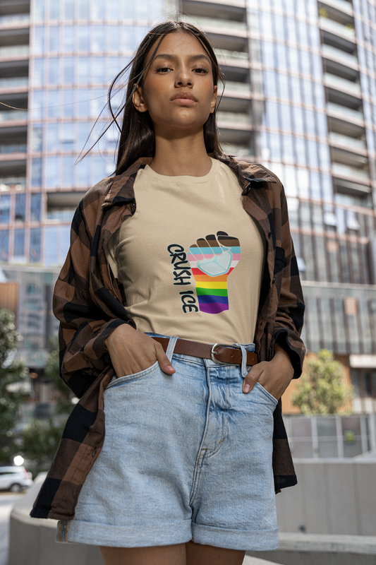 An Indigenous woman stands confidently against a modern city building. She wears a cream-colored, slim-fit t-shirt featuring the Crush ICE Rainbow Progress design—a protest fist shattering ice, filled with horizontal rainbow and brown/black/trans-inclusive stripes. Her tee is tucked into light-wash denim shorts with a brown belt, highlighting the fitted silhouette. She wears an open brown-and-black plaid flannel over her shoulders, embodying urban community resistance.