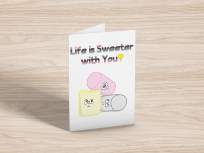 A greeting card on light wood background. The card has a white background. At the top is the text, "Life is Sweeter with You!" in the queerplatonic pride flag colours. Below are matching pink, yellow, and grey cartoon marshmallows with cute expressions. 